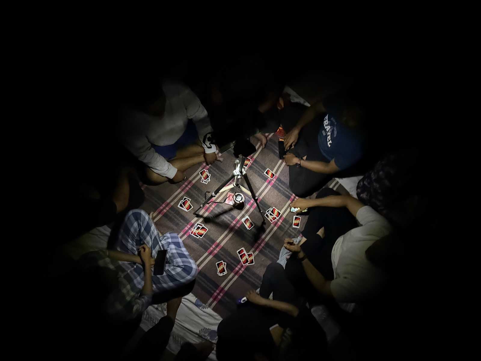 A group of friends and colleagues are gathered in a circle on a mat, playing a game of Uno under the dim light of a portable lamp or flashlight during their overnight camping trip at Savlya Ghat.