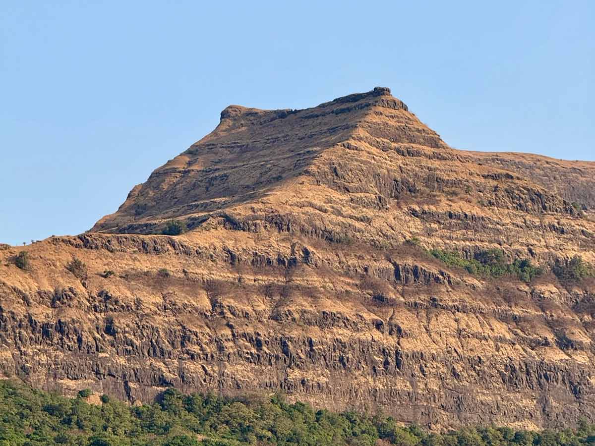 A prominent brown, rugged hill, likely Jambhya Dongar as seen from Savlya Ghat, with a distinctive flat top and tiered rock formations below, standing out against a clear blue sky.