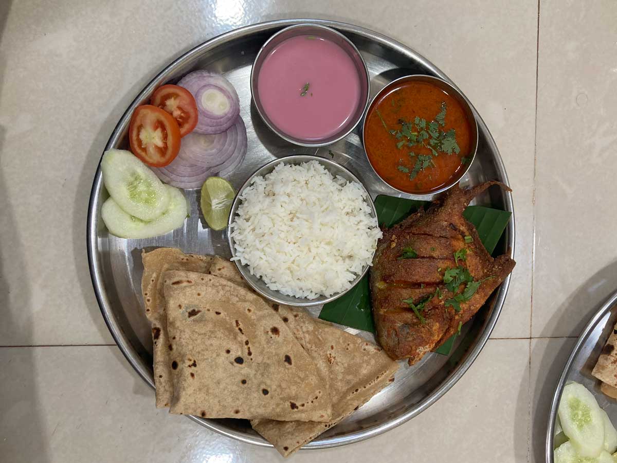 Traditional Konkan fish thali served on a steel plate, featuring fried fish, curry, solkadhi (pink coconut-based drink), rice, bhakri, fresh salad with cucumber, tomato, onion, and lime
