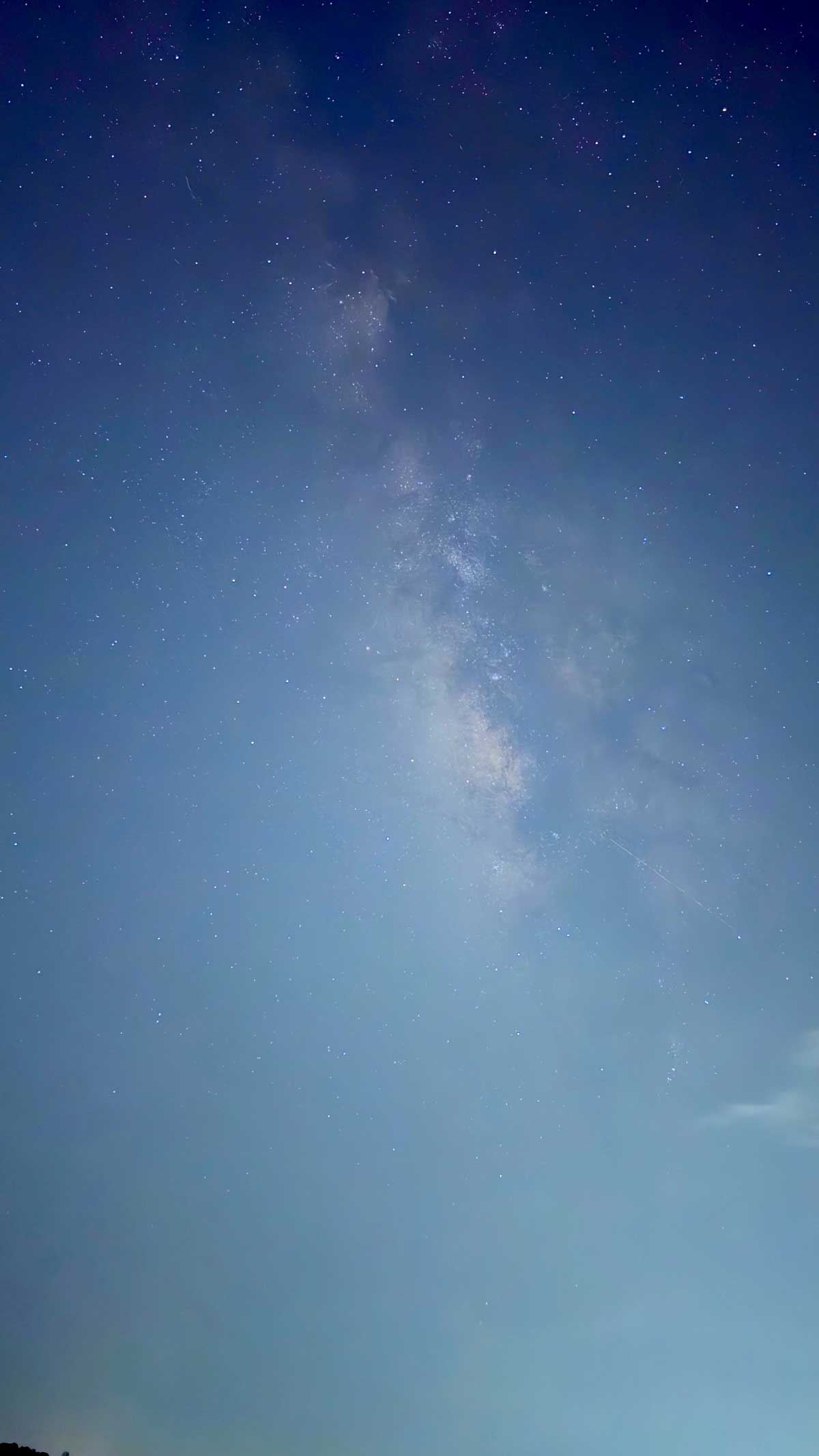 A vertical shot of the night sky revealing a hazy but distinct band of the Milky Way stretching across the frame, with countless stars visible against a deep blue to lighter blue gradient, taken during a challenging but successful attempt at astrophotography from Savlya Ghat.
