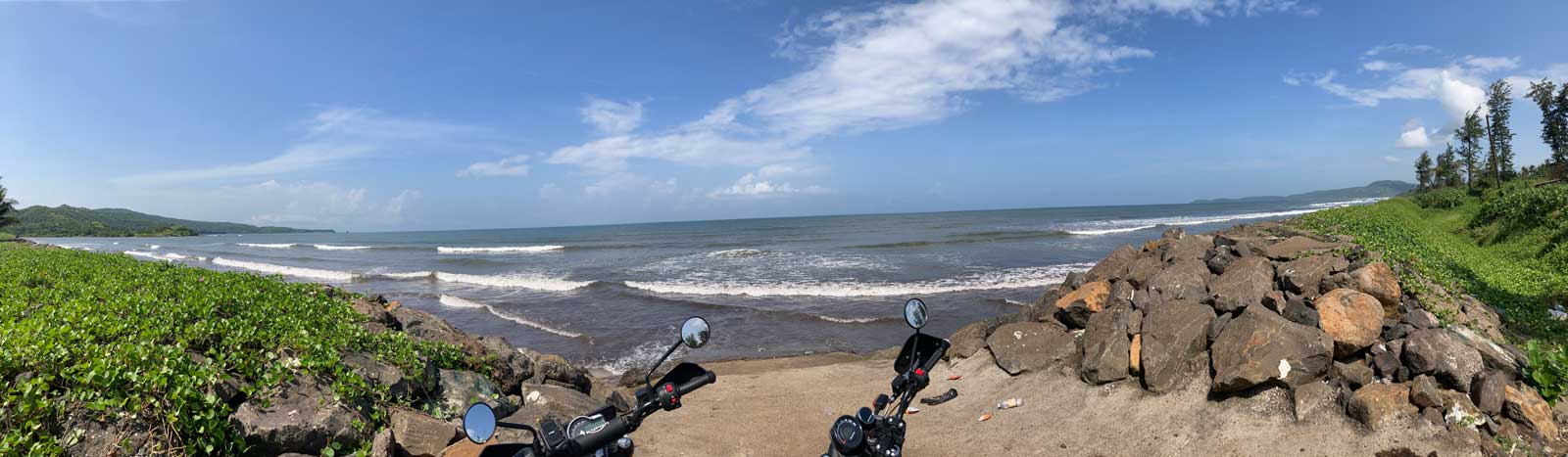 Panoramic view of Diveagar Beach showing two motorcycles (Triumph Scrambler 400X and RE Scram 411) parked at the shoreline with waves breaking and coconut trees lining the coast