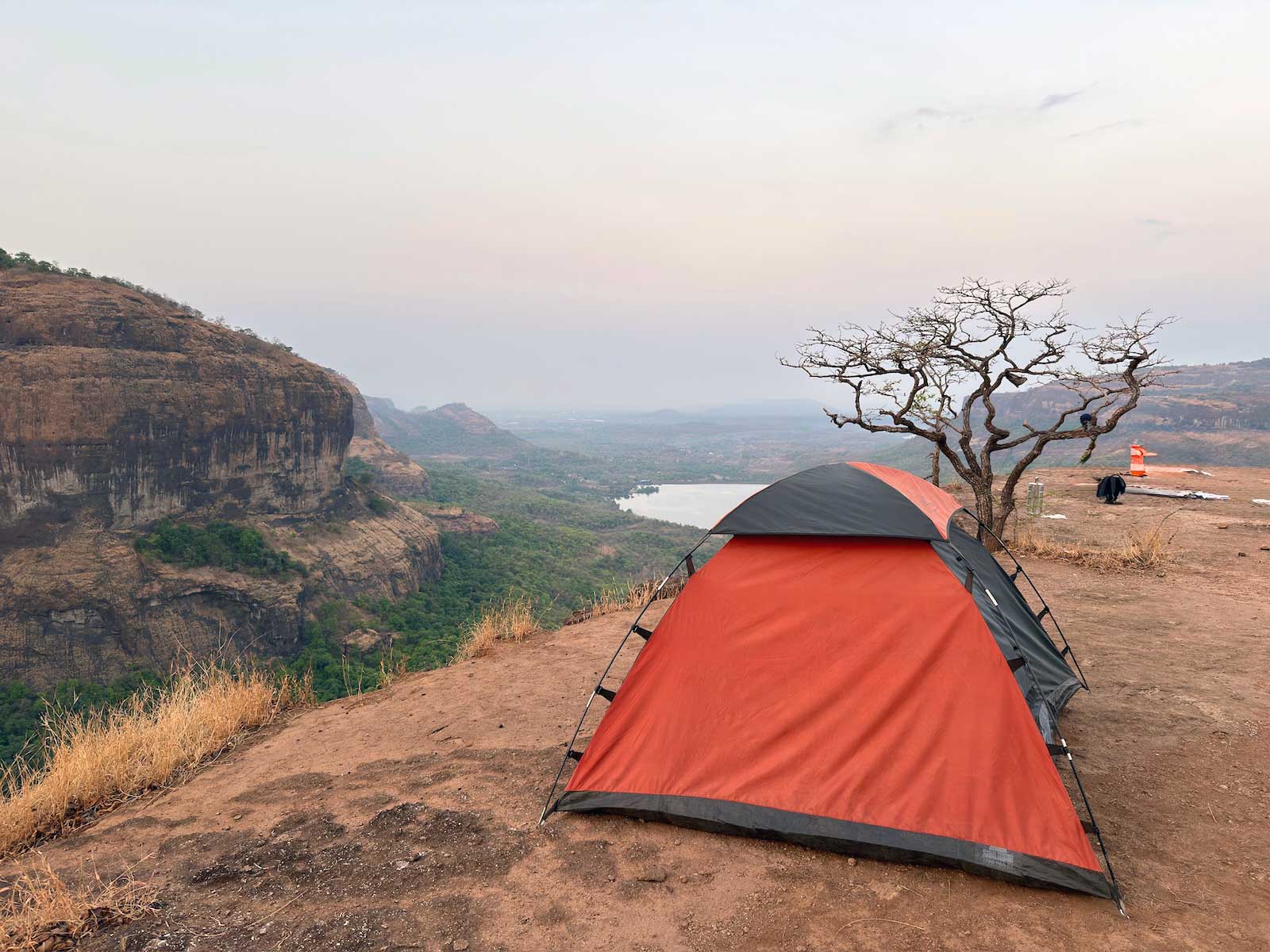 A vibrant orange and grey camping tent pitched on a reddish-brown plateau at Savlya Ghat, overlooking a deep valley with lush green patches and the distant backwaters of the Bhira Dam under a cloudy sky. A gnarled, bare tree stands beside the tent.
