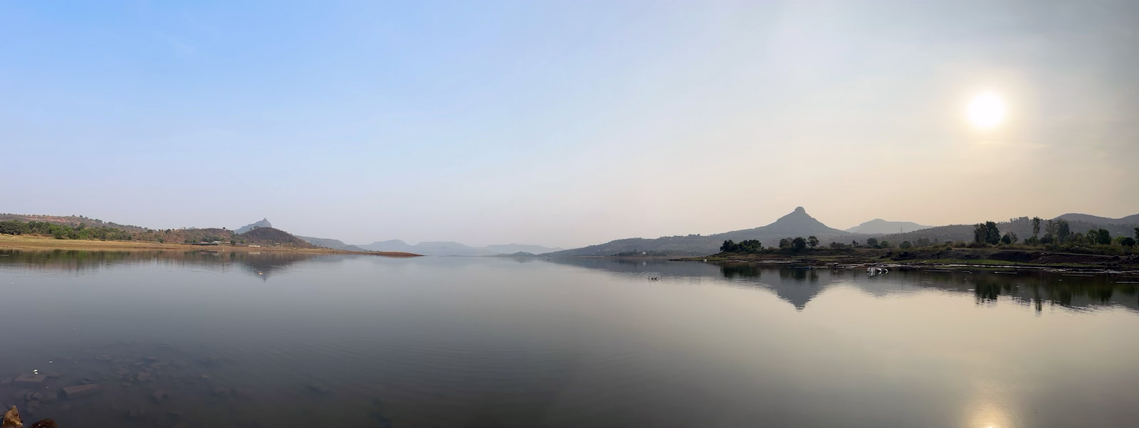 Wide panoramic view of tranquil Pawana Lake at dawn with perfect reflections, featuring Tikona Fort visible on the left and the distant silhouettes of Lohgad and Visapur forts rising against the hazy morning sky along the Sahyadri mountain range