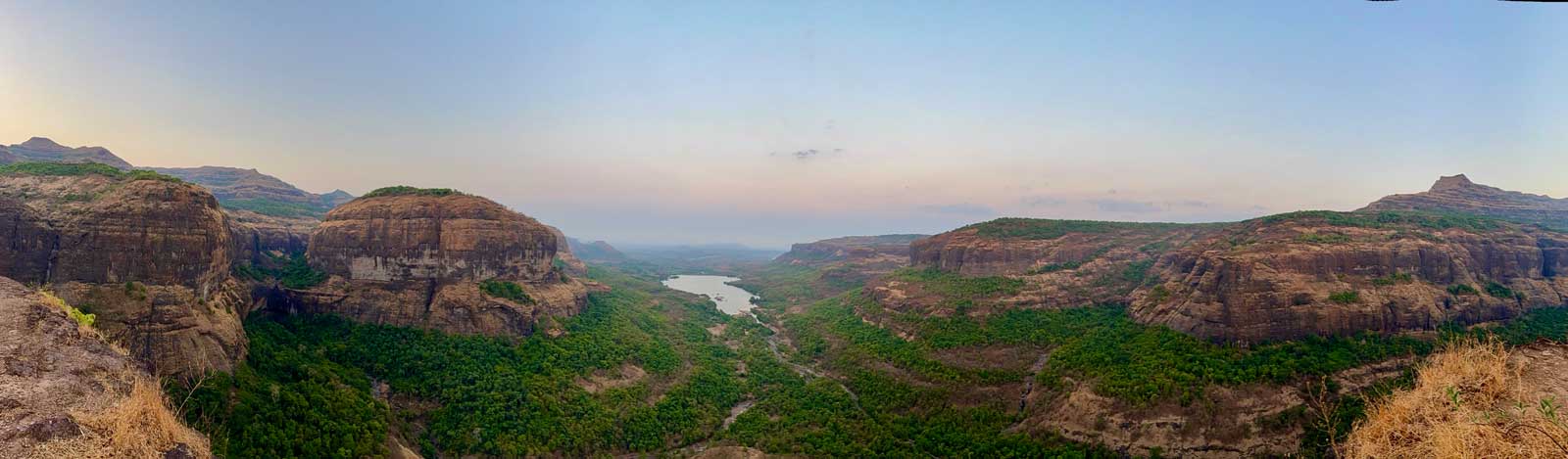 A wide panoramic shot at dusk shows the vast landscape of Savlya Ghat, with rocky, flat-topped hills characteristic of the Deccan Trap, lush green valleys, and the shimmering backwaters of the Bhira Dam nestled below.