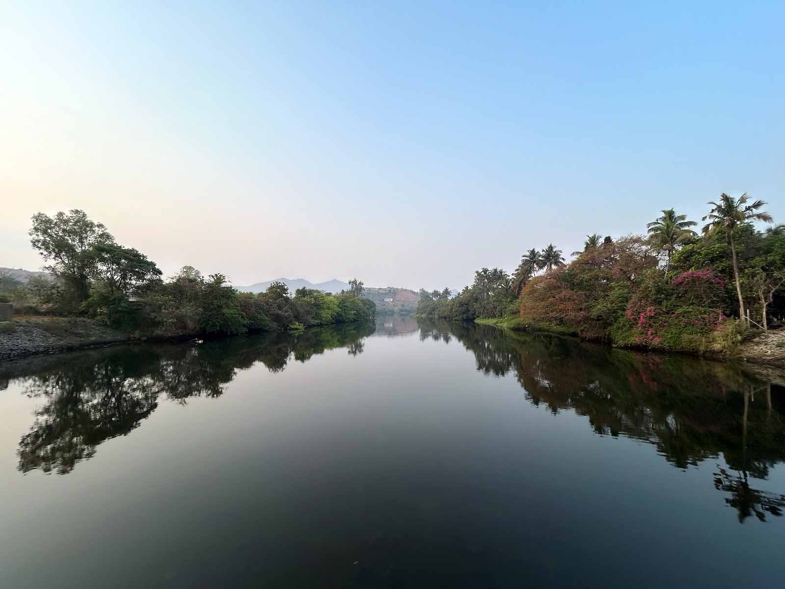 Serene morning view of Pawana River from Kadadhe Bridge, showing perfect reflections of trees, palm groves and distant mountains in the still water, captured during the early morning motorcycle ride stop