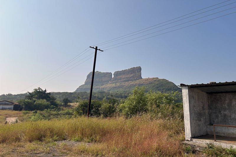 Panoramic view of Tailbaila Fort perched on a hill as seen from the parking area, with its weathered stone ramparts standing against the backdrop of the expansive Kundalika Valley and surrounding mountain ranges
