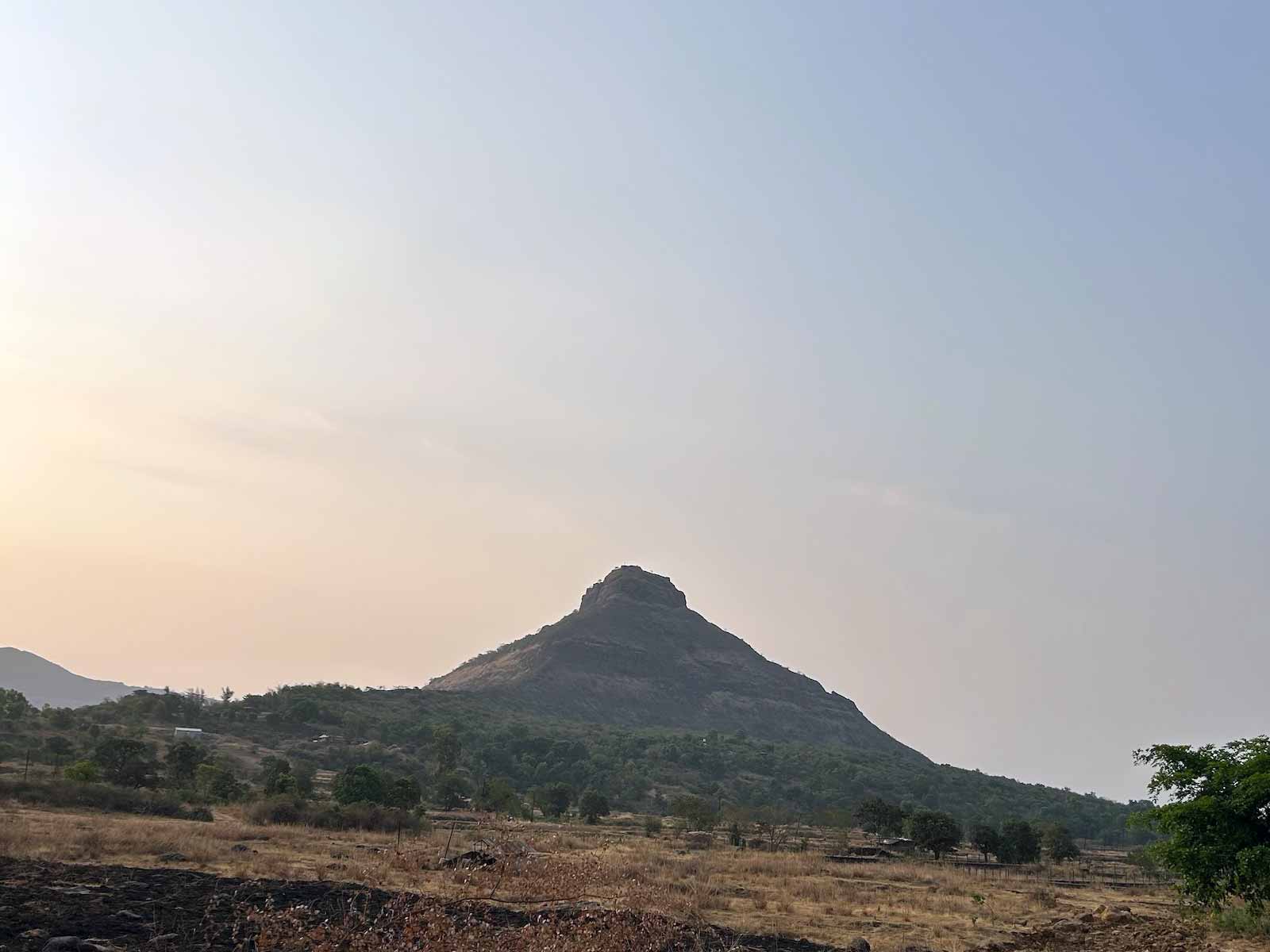 The distinctive triangular silhouette of Tikona Fort rising dramatically against the morning sky, visible along the motorcycle route to Wagheshwar Temple