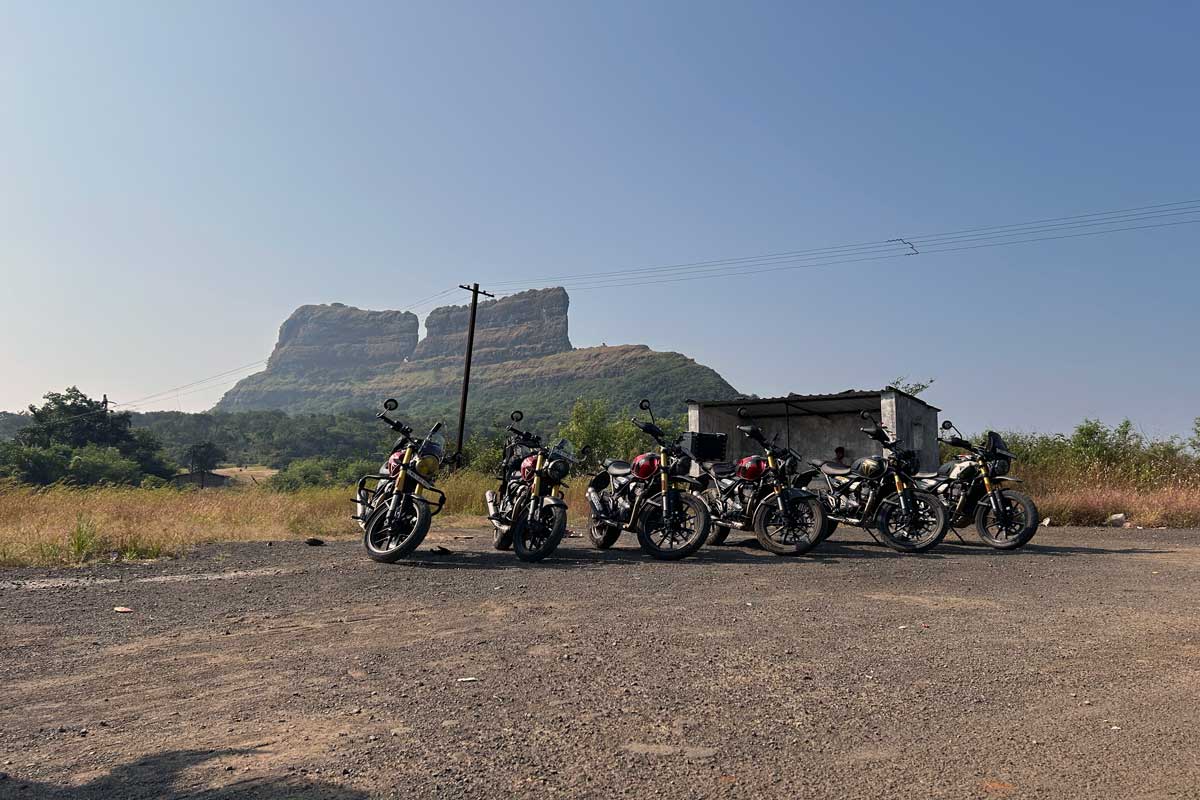 Six Triumph motorcycles (three Speed 400s and three Scrambler 400X models) lined up in front of the ancient stone walls of Tailabaila Fort, with verdant hills and valleys of the Western Ghats visible in the background